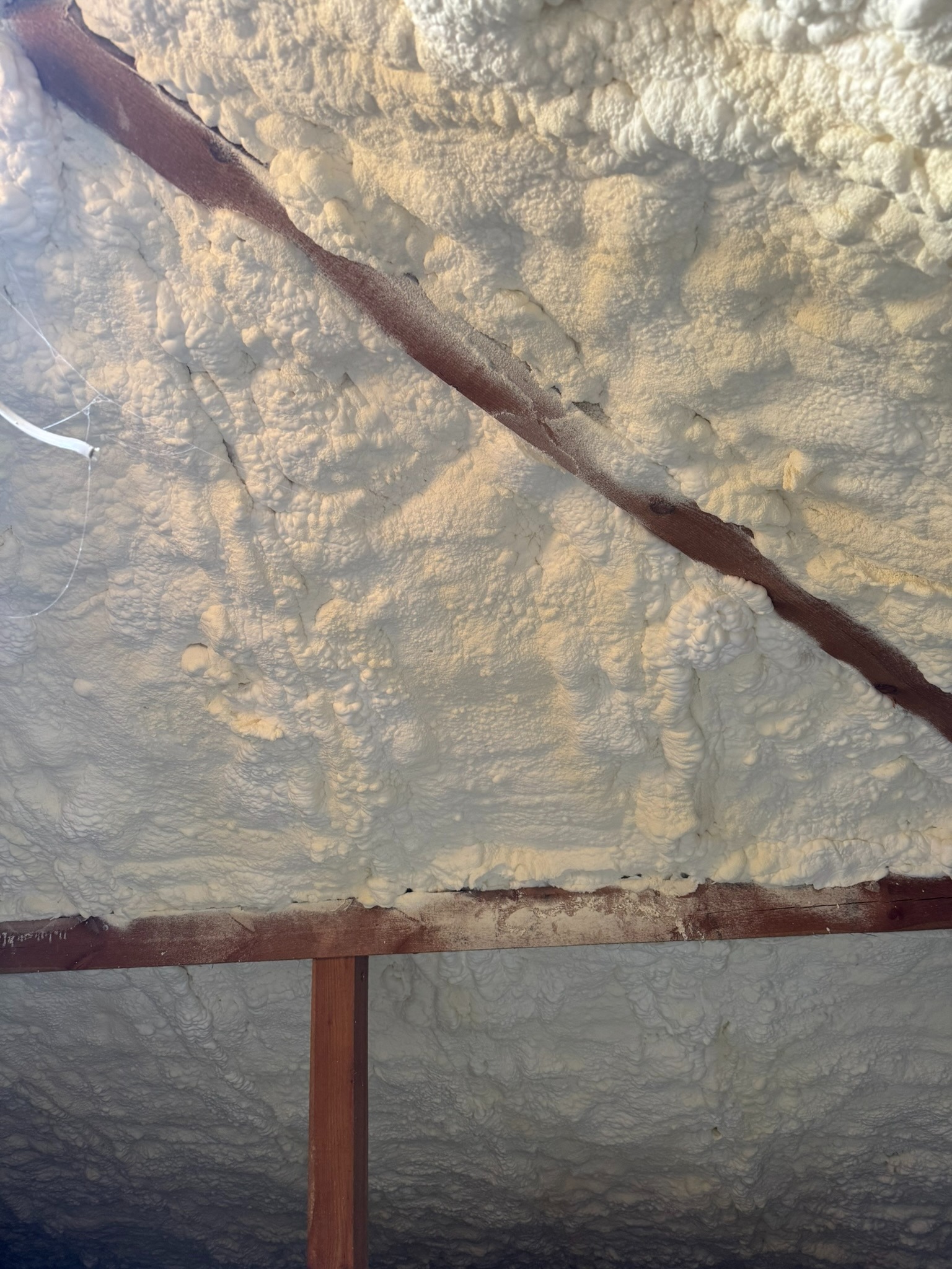 Ocieplenie poddasza pianką PUR, widoczne drewniane elementy konstrukcyjne. Biała, nierówna powierzchnia pianki wypełnia przestrzeń między belkami.