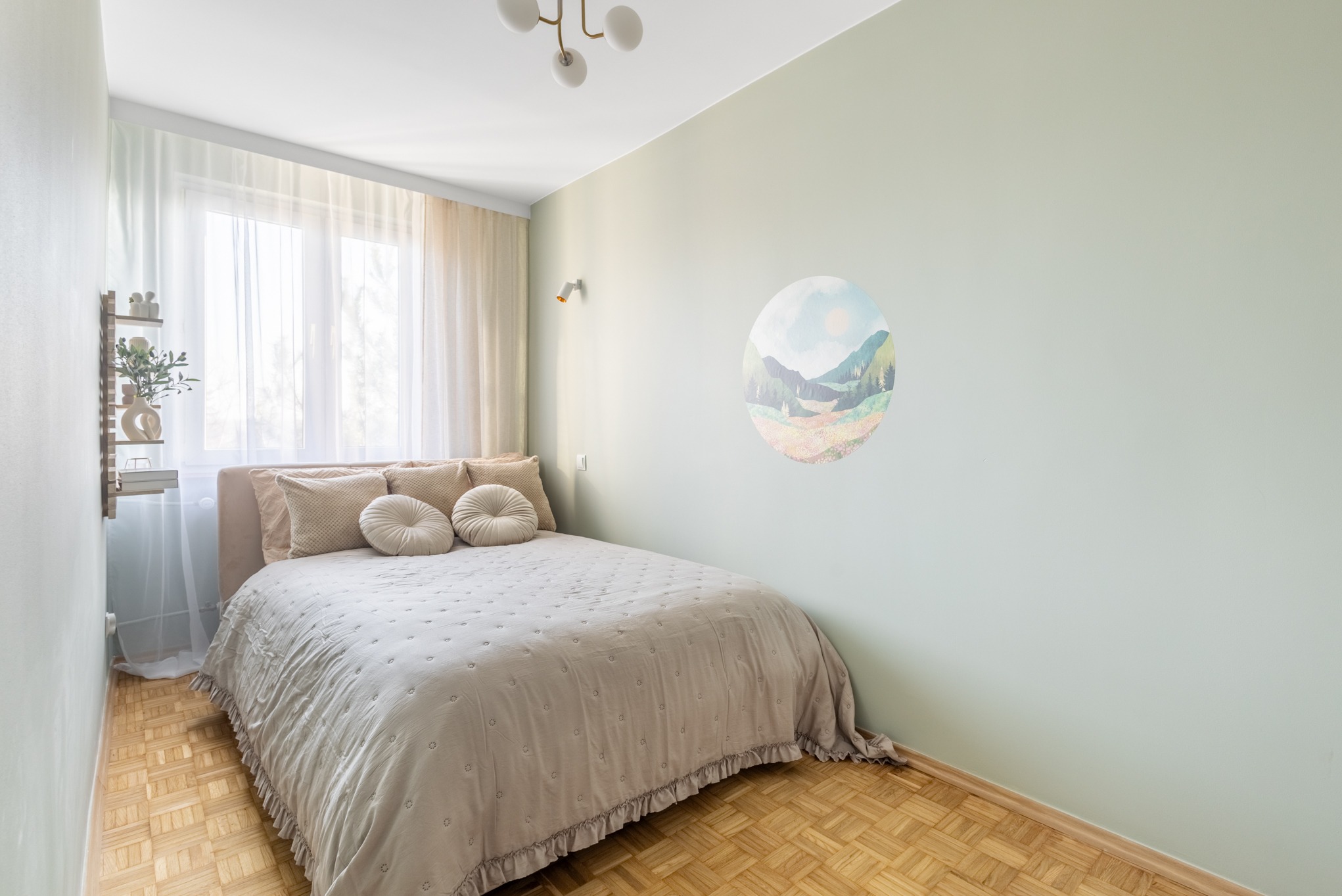 Jasna sypialnia z łóżkiem, drewnianą podłogą i okrągłą naklejką z motywem górskim na ścianie. Minimalistyczny wystrój, naturalne światło.