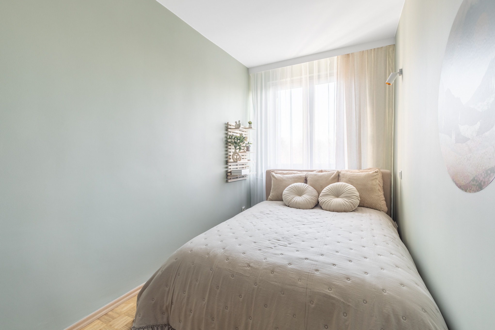 Jasna sypialnia w odcieniach beżu i zieleni: łóżko z okrągłymi poduszkami, drewniana półka z roślinami, okno z firanką i okrągła dekoracja na ścianie.