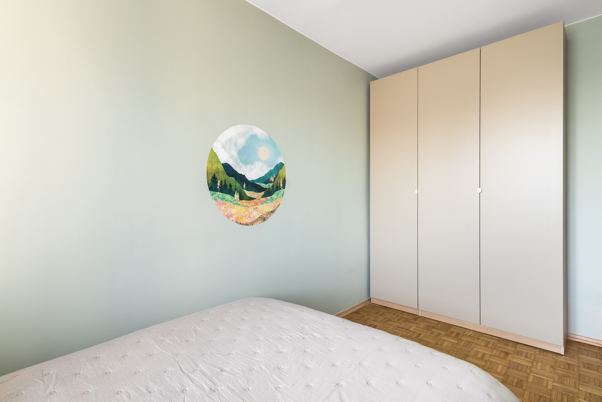 Jasny pokój z łóżkiem, parkietem i beżową szafą. Na ścianie okrągła dekoracja z motywem górskiego krajobrazu. Minimalistyczny design wnętrza.