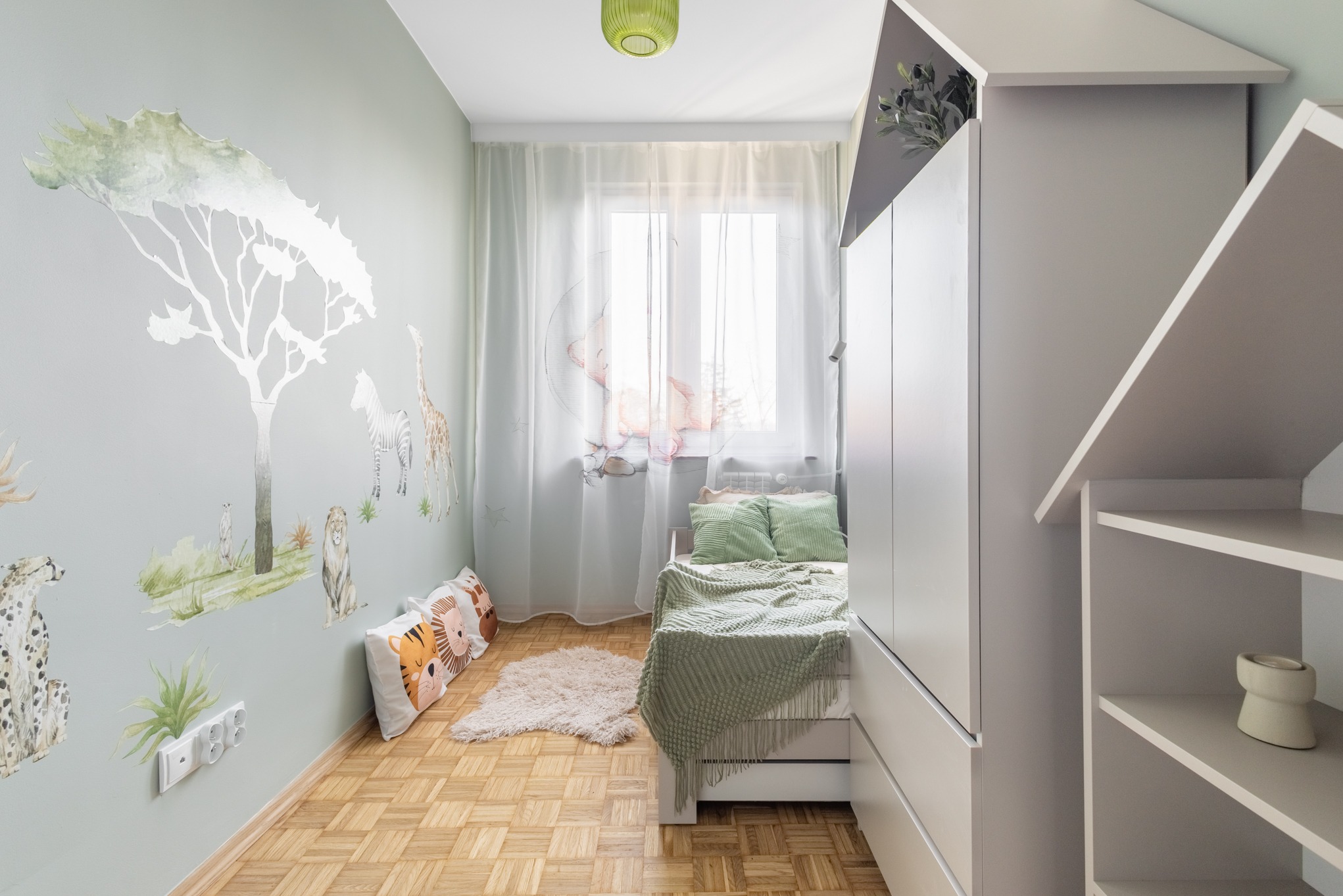 Pokój dziecięcy z motywem safari: malowidła zwierząt na ścianie, drewniana podłoga, białe meble i okno z delikatnymi zasłonami. Przytulna aranżacja.