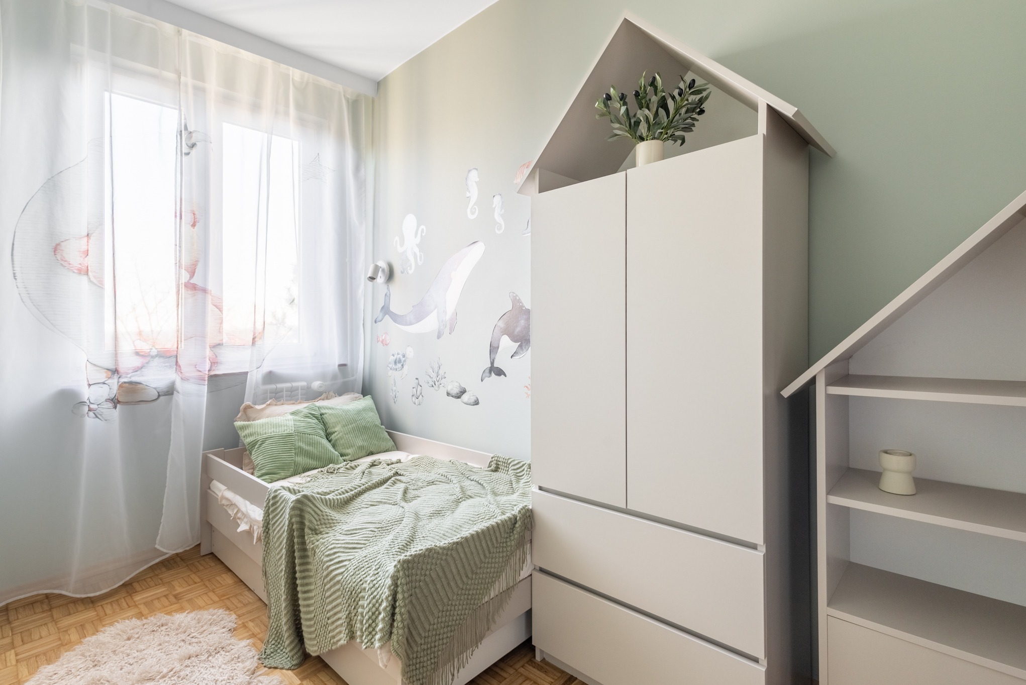Pokój dziecięcy w pastelowych barwach z motywem morskim na ścianie, białe meble w kształcie domków, łóżko z zielonym pledem i poduszkami, jasne zasłony.