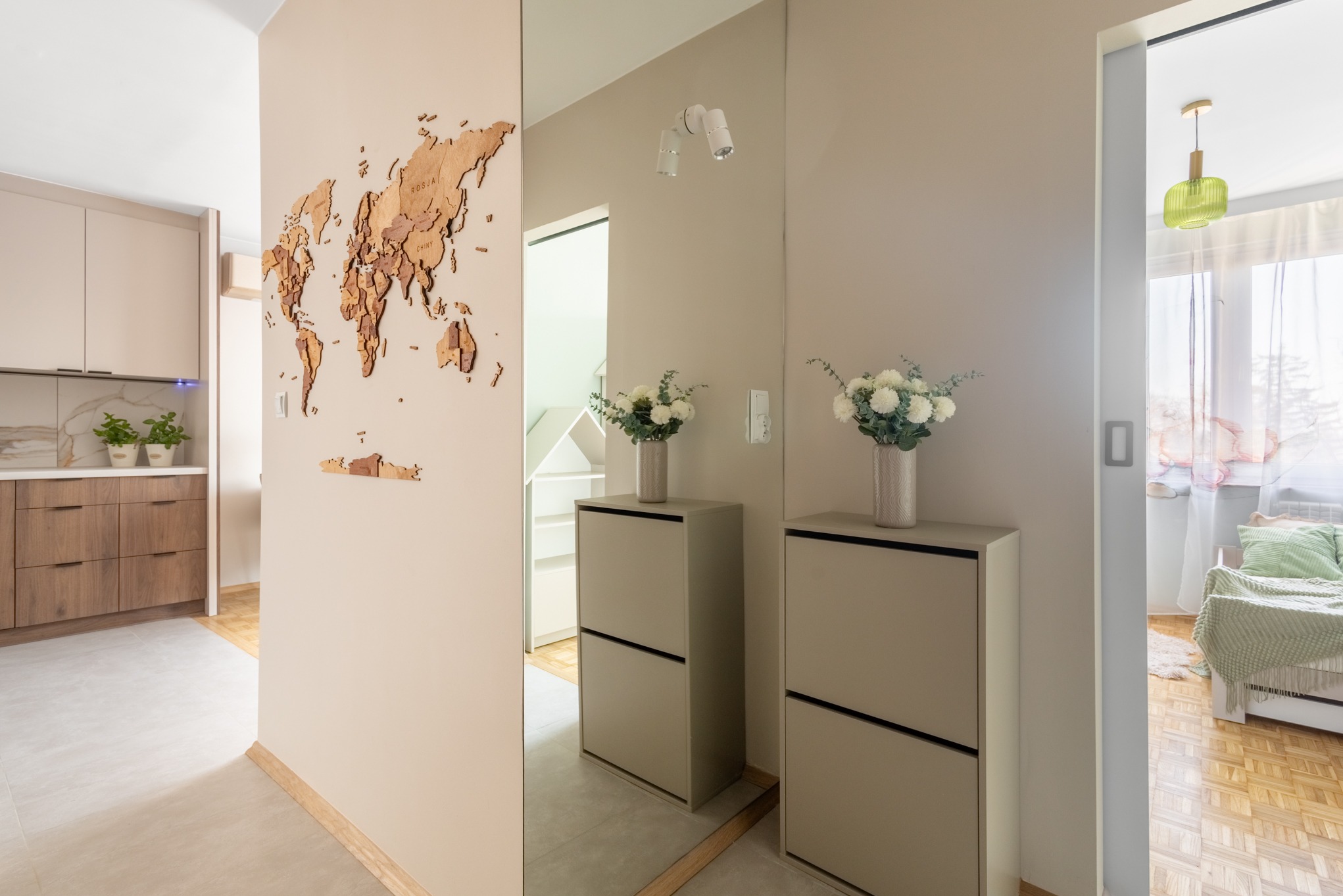 Jasne wnętrze mieszkania z drewnianą mapą świata na ścianie, lustrem odbijającym korytarz i widokiem na pokój przez przesuwne drzwi.