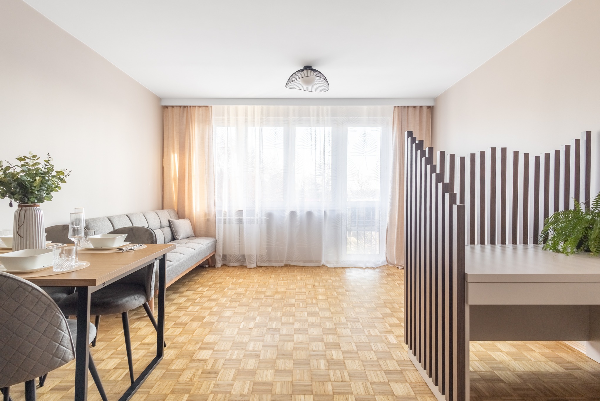 Jasne wnętrze pokoju dziennego z parkietem w jodełkę, szarą sofą i stołem z nakryciem. Dekoracyjna ścianka działowa z pionowych listew dodaje charakteru.