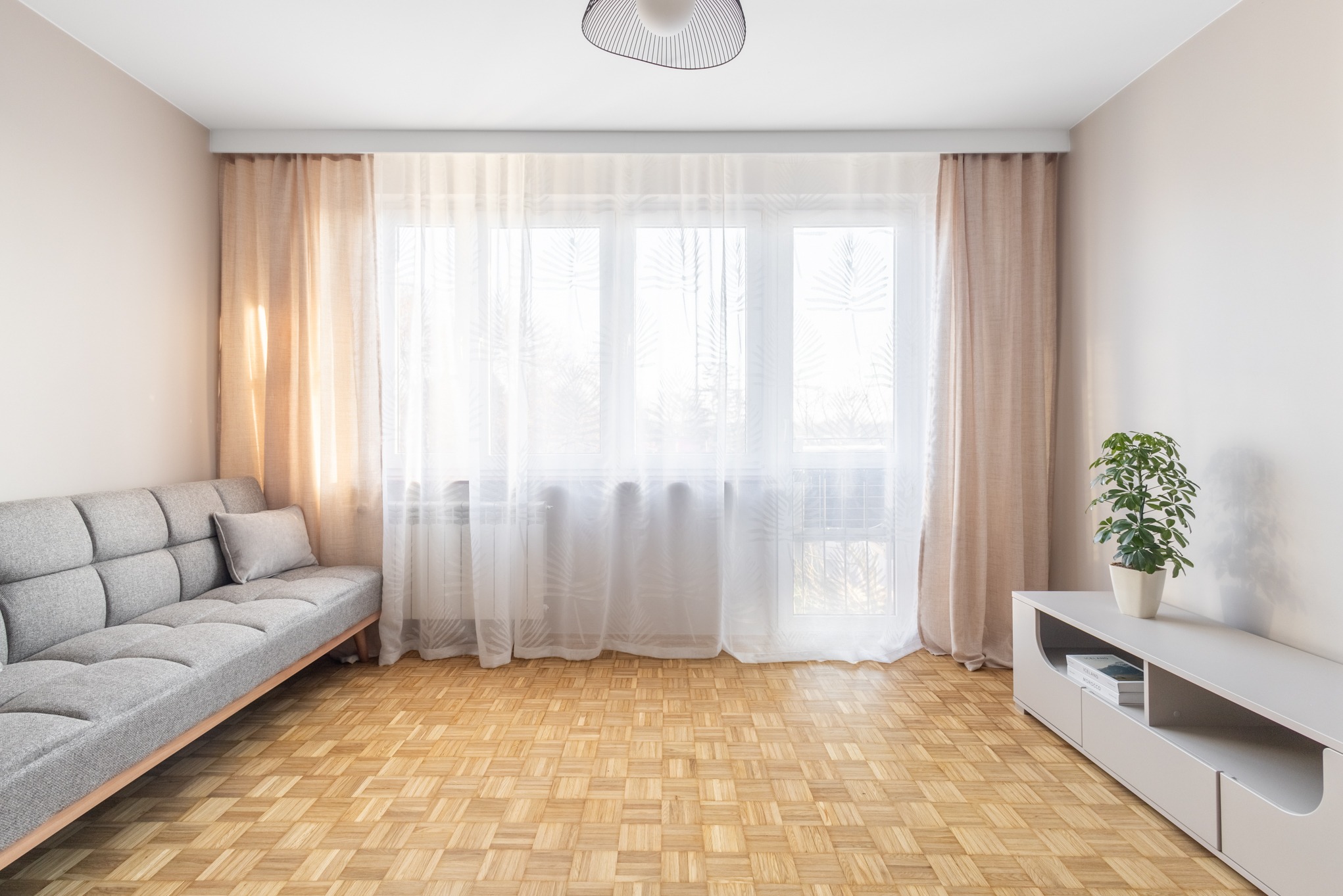 Jasne wnętrze salonu z parkietem w jodełkę, szarą sofą i białymi zasłonami. Widoczne okno z delikatnym wzorem, lampa sufitowa i roślina doniczkowa na szafce RTV.