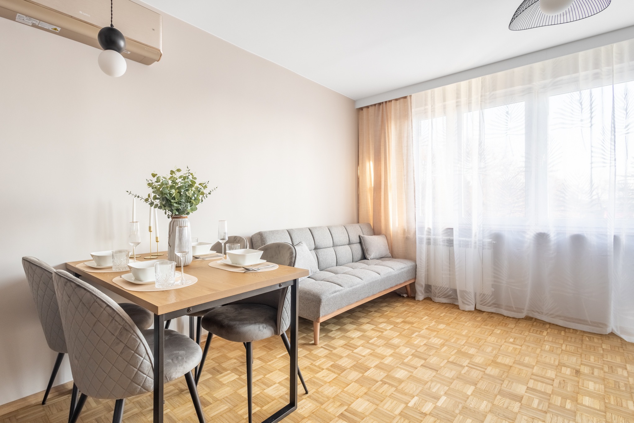 Jasne wnętrze pokoju dziennego z drewnianą podłogą, szarą sofą i stołem z nakryciem. Minimalistyczny design, naturalne światło wpadające przez okno.