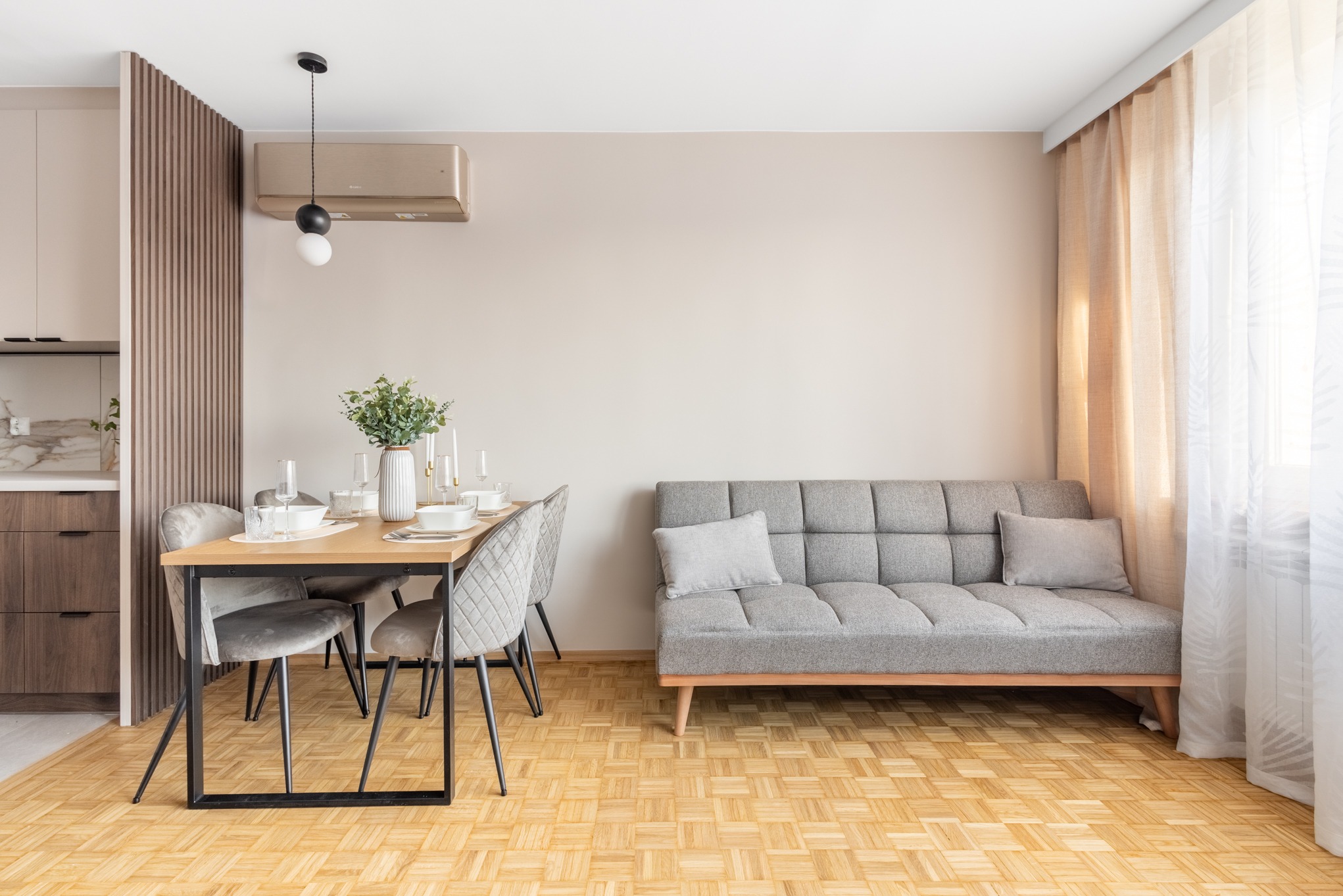 Jasne wnętrze: parkiet w jodełkę, szara sofa, stół z zastawą, klimatyzator i designerska lampa wisząca. Minimalistyczny design z elementami drewna i beżu.
