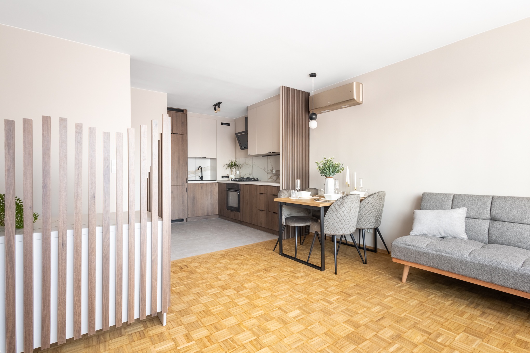 Jasne, przestronne wnętrze po remoncie: kuchnia z jadalnią i salonem. Parkiet w jodełkę, drewniane lamele, szara sofa i stół z krzesłami. Nowoczesny design.