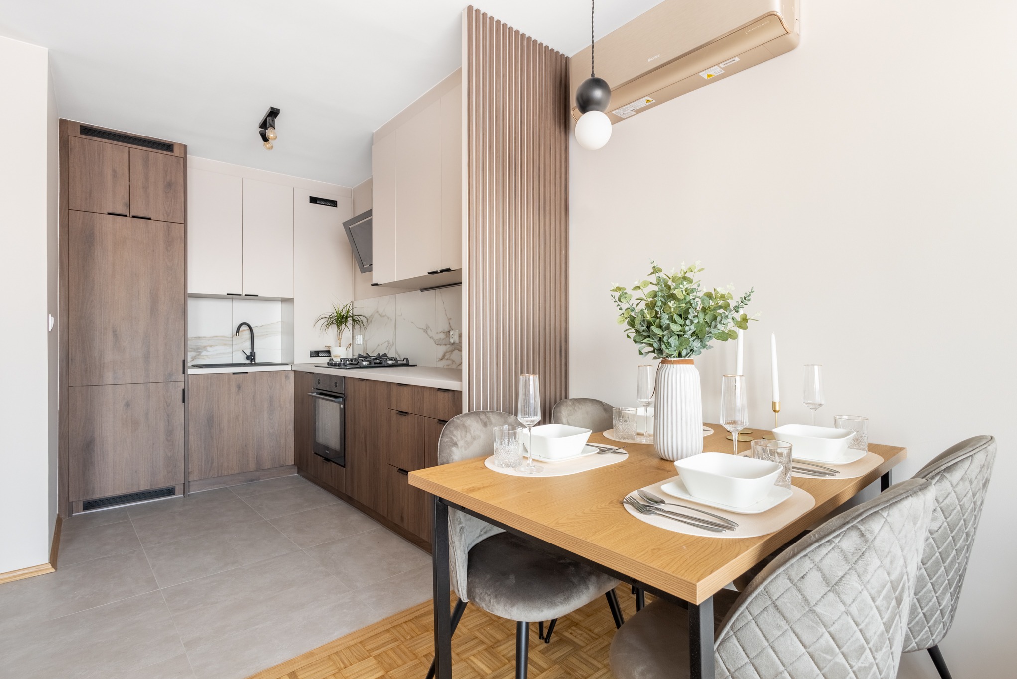 Nowoczesna kuchnia z jadalnią: drewniane szafki, marmurowy backsplash, stół z nakryciem i szare krzesła. Minimalistyczny design, jasne wnętrze z akcentami drewna.