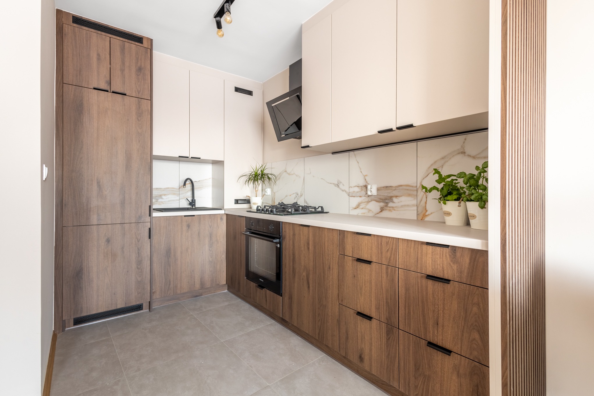 Nowoczesna kuchnia w beżach i brązach: zabudowa meblowa z drewnianymi frontami, marmurowy backsplash, czarne akcenty i rośliny doniczkowe. Minimalistyczny design.