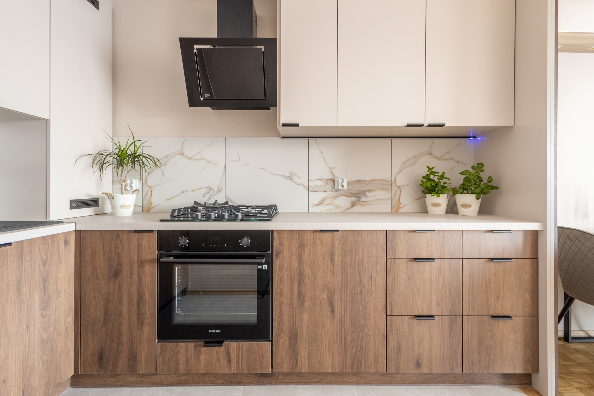 Nowoczesna kuchnia z drewnianymi szafkami, marmurowym backsplashiem i czarnym okapem. Rośliny w doniczkach dodają świeżości. Minimalistyczny design z dbałością o detale.