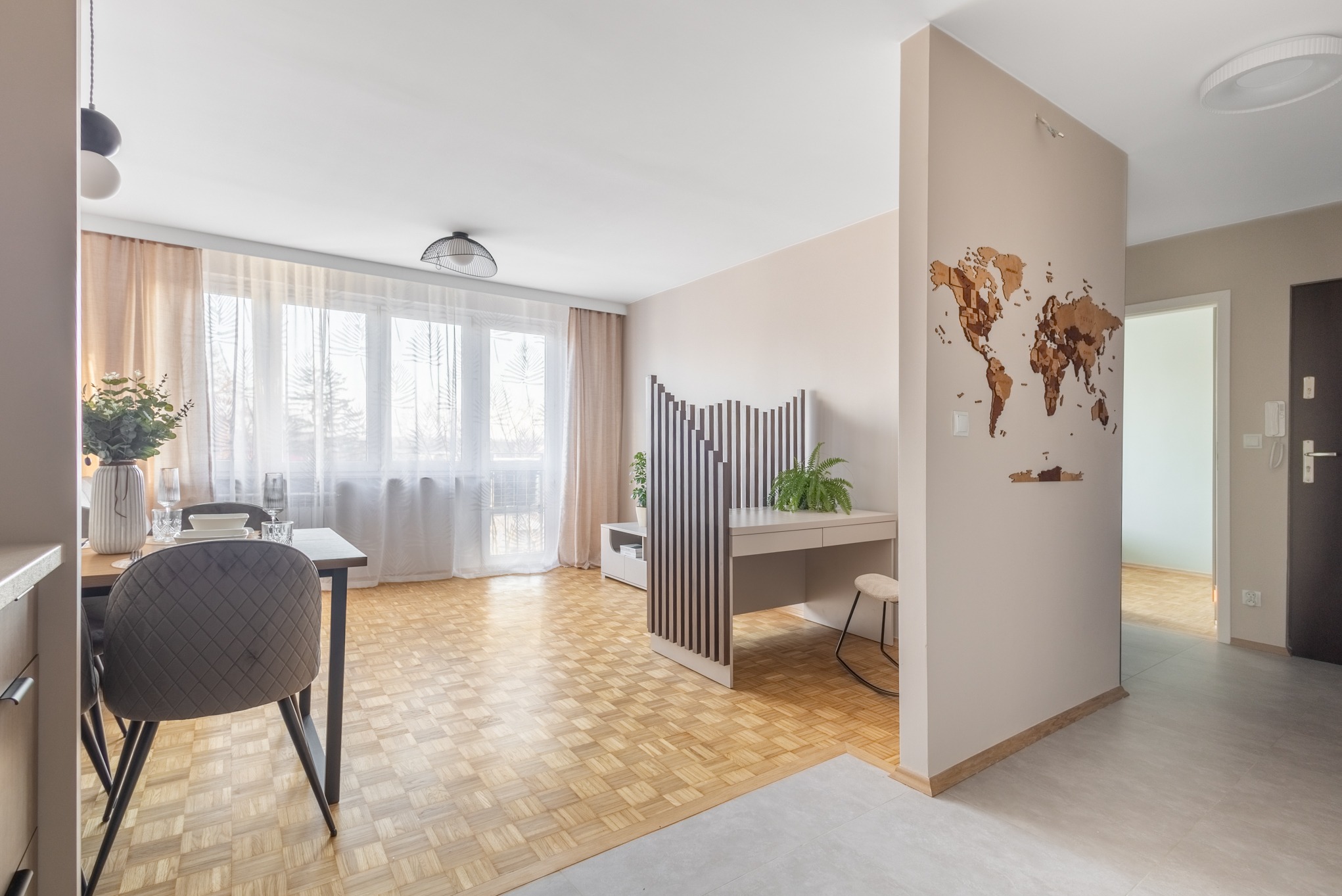 Jasne wnętrze mieszkania z parkietem w jodełkę, biurkiem z ażurową ścianką działową i dekoracją ścienną w postaci mapy świata. Widok na jadalnię i okno.