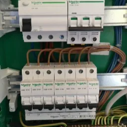 Rozdzielnica elektryczna z zamontowanymi bezpiecznikami i przewodami w zielonej obudowie, widoczne elementy marki Schneider Electric.