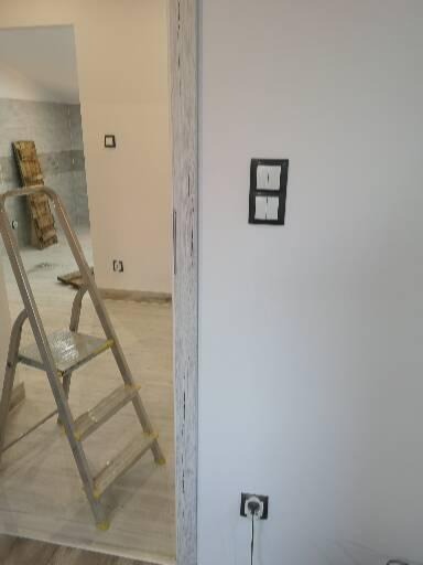 Biała ściana z zamontowanym podwójnym włącznikiem światła w czarnej ramce i gniazdkiem elektrycznym, drabina rozstawiona w pomieszczeniu obok, widok z bliska.