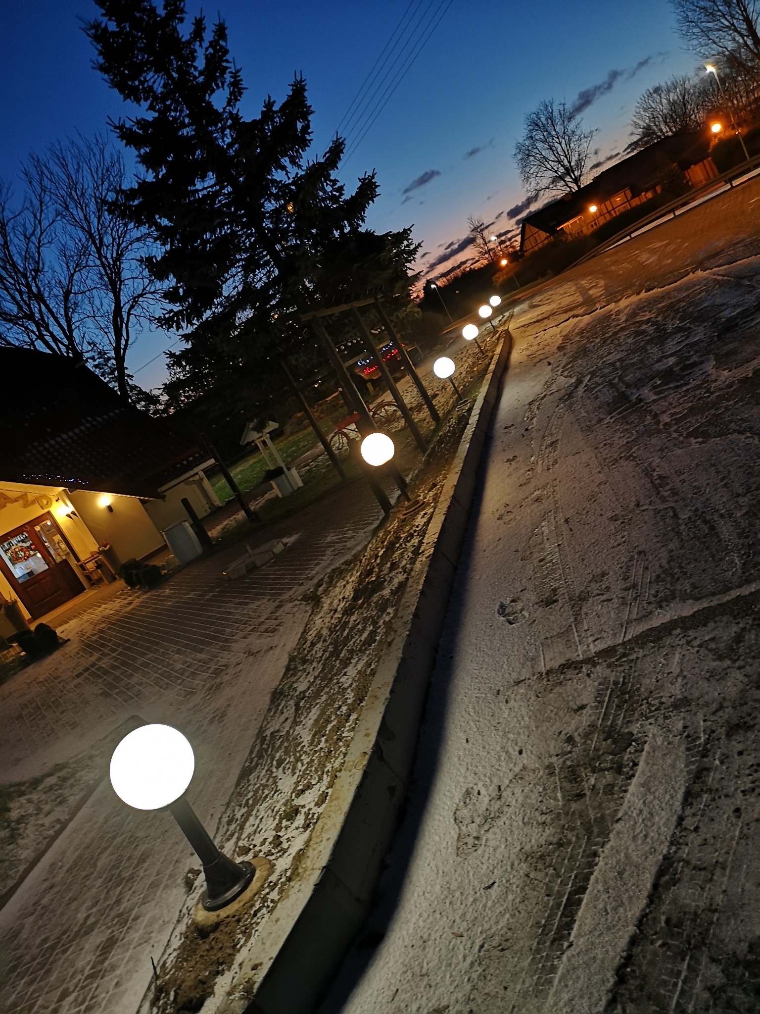 Oświetlona ścieżka z kulistymi lampami wzdłuż budynku w zimowej scenerii o zmierzchu, widoczne ślady opon na śniegu.