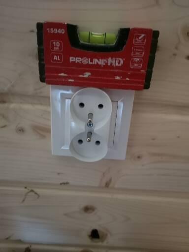 Białe gniazdko elektryczne z bolcem uziemiającym, sprawdzane poziomicą Proline HD na drewnianej ścianie z boazerii.
