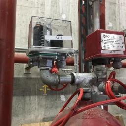 H.V.E - Instalacja alarmowa z czujnikiem przepływu w systemie rur, widoczna w Gorlicach. Czerwone rury i przewody oraz szara betonowa ściana w tle. Na czujniku widoczna etykieta A1-2.