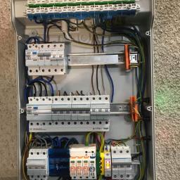 H.V.E - Otwarta skrzynka bezpiecznikowa z zainstalowanymi bezpiecznikami, przewodami i listwami zaciskowymi na szarej ścianie o chropowatej fakturze.