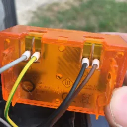 Pomarańczowa obudowa lampy ostrzegawczej z podłączonymi przewodami: zielono-żółtym, szarym, czarnym i brązowym, widoczne oznaczenia CE i adres producenta.