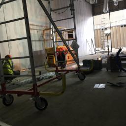 H.V.E - Wnętrze w trakcie prac instalacyjnych: widoczne rusztowania, podnośnik, pracownicy w kamizelkach odblaskowych, instalacje pod sufitem, narzędzia i materiały budowlane.