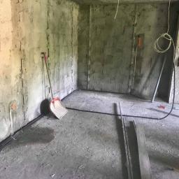 H.V.E - Surowe wnętrze w trakcie prac wykończeniowych: widoczne przewody elektryczne przy ścianie, szpachlowane ściany z widocznymi bruzdami, łopata oparta o ścianę, lampa budowlana na statywie i fragment...