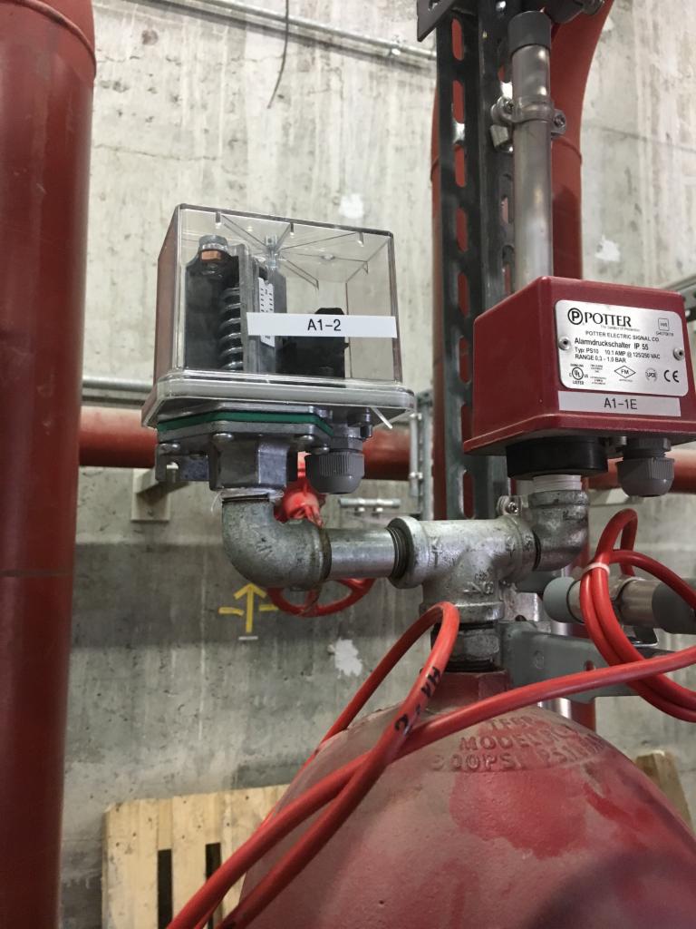 Instalacja alarmowa z czujnikiem przepływu w systemie rur, widoczna w Gorlicach. Czerwone rury i przewody oraz szara betonowa ściana w tle. Na czujniku widoczna etykieta A1-2.