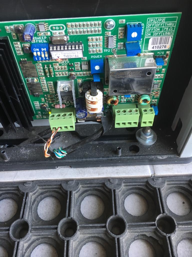 Otwarta obudowa urządzenia elektronicznego z widoczną zieloną płytką drukowaną, elementami elektronicznymi, złączami śrubowymi i przewodami. Widoczne ustawienia JP1 i JP2. Obok leży gumowa mata.