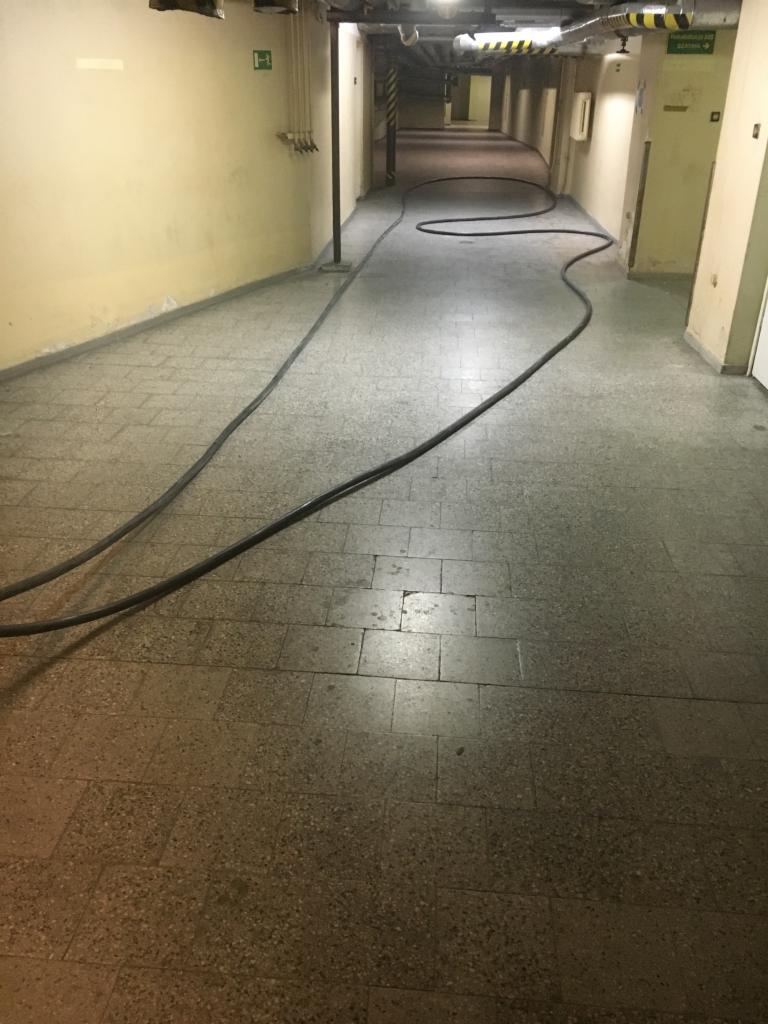 Długi, czarny kabel elektryczny wijący się przez pusty korytarz z żółtymi ścianami i szarą podłogą z płytek. Kabel biegnie wzdłuż ściany i skręca w głąb korytarza.