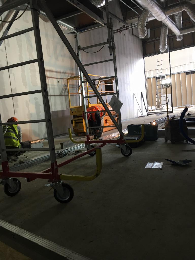 Wnętrze w trakcie prac instalacyjnych: widoczne rusztowania, podnośnik, pracownicy w kamizelkach odblaskowych, instalacje pod sufitem, narzędzia i materiały budowlane.