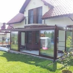 Almont System Fabryka Ogrodów Zimowych - Szklana zabudowa tarasu z brązowymi ramami przylegająca do domu z balkonem, widoczne elementy montażowe i pracownicy w tle.