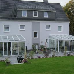 Almont System Fabryka Ogrodów Zimowych - Dwa przeszklone ogrody zimowe z białymi ramami, przylegające do szarego domu z ciemnym dachem, widok z trawnika.