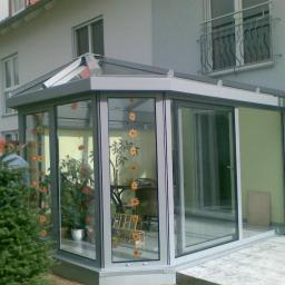 Almont System Fabryka Ogrodów Zimowych - Szklana oranżeria z aluminiową ramą przylegająca do domu, ozdobiona naklejkami w kształcie słoneczników, z widocznymi roślinami w doniczkach wewnątrz.