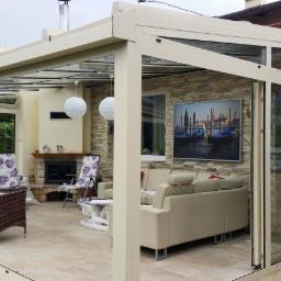 Almont System Fabryka Ogrodów Zimowych - Zadaszona przestrzeń tarasowa z przesuwnymi szklanymi drzwiami, beżowa rama, kamienna ściana z obrazem weneckich gondoli, jasny narożnik i kominek w tle.