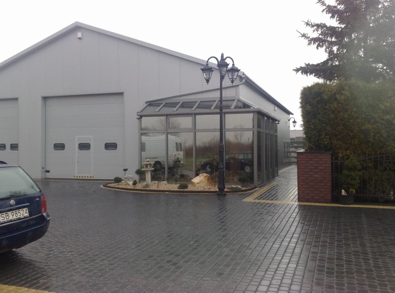 Szklana konstrukcja przylegająca do hali magazynowej, oświetlona ozdobną latarnią, z widocznym fragmentem parkingu z kostki brukowej i zaparkowanym samochodem osobowym.