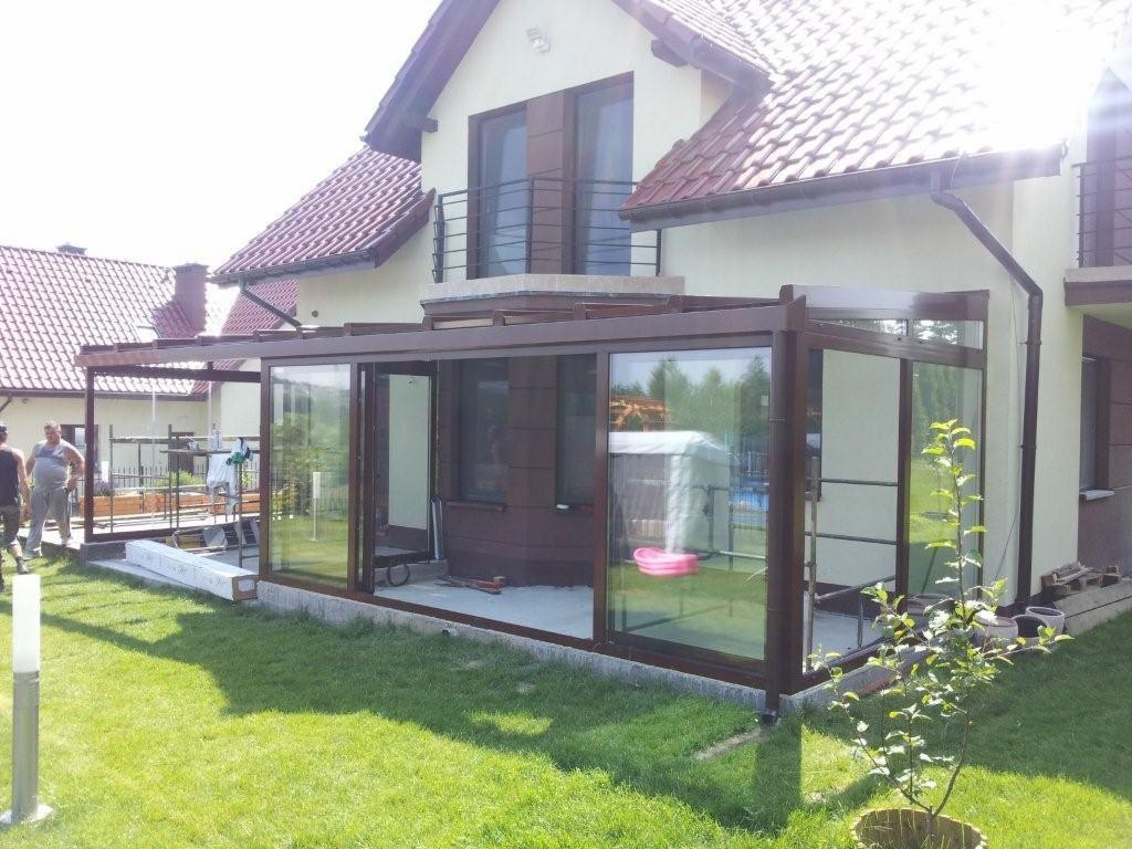 Szklana zabudowa tarasu z brązowymi ramami przylegająca do domu z balkonem, widoczne elementy montażowe i pracownicy w tle.