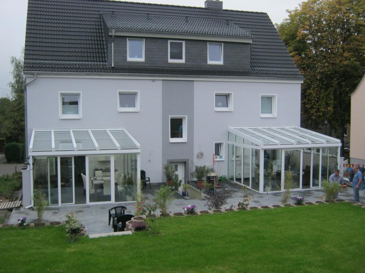 Dwa przeszklone ogrody zimowe z białymi ramami, przylegające do szarego domu z ciemnym dachem, widok z trawnika.