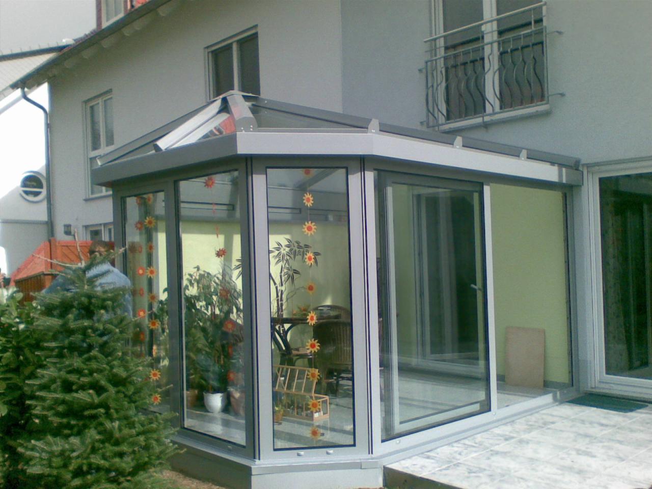 Szklana oranżeria z aluminiową ramą przylegająca do domu, ozdobiona naklejkami w kształcie słoneczników, z widocznymi roślinami w doniczkach wewnątrz.