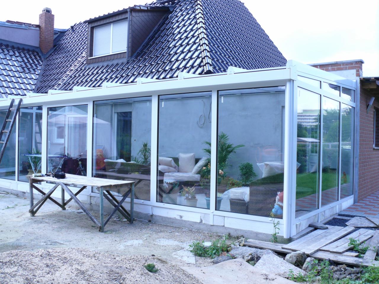 Szklana zabudowa tarasu przylegająca do domu z widocznym wnętrzem z meblami ogrodowymi, na zewnątrz stół roboczy i drabina.