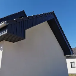 Nowoczesny dom jednorodzinny z grafitowym dachem z blachy na rąbek stojący i orynnowaniem, widok z dołu na fragment elewacji i dach w słoneczny dzień.