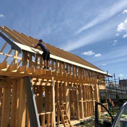 Nord-Haus-Inwest - Mężczyzna mocuje płytę do drewnianej konstrukcji dachu domu w trakcie budowy, widok z dołu na tle błękitnego nieba.