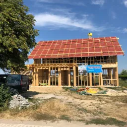 Wznoszenie konstrukcji szkieletowej domu w Powałkach z widoczną wstępną warstwą dachu w kolorze czerwonym i banerem reklamowym na elewacji, na tle błękitnego nieba.