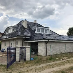 Dom z jasnej cegły z nowym, ciemnym dachem z dachówki ceramicznej, widok z zewnątrz, błękitna brama wjazdowa.