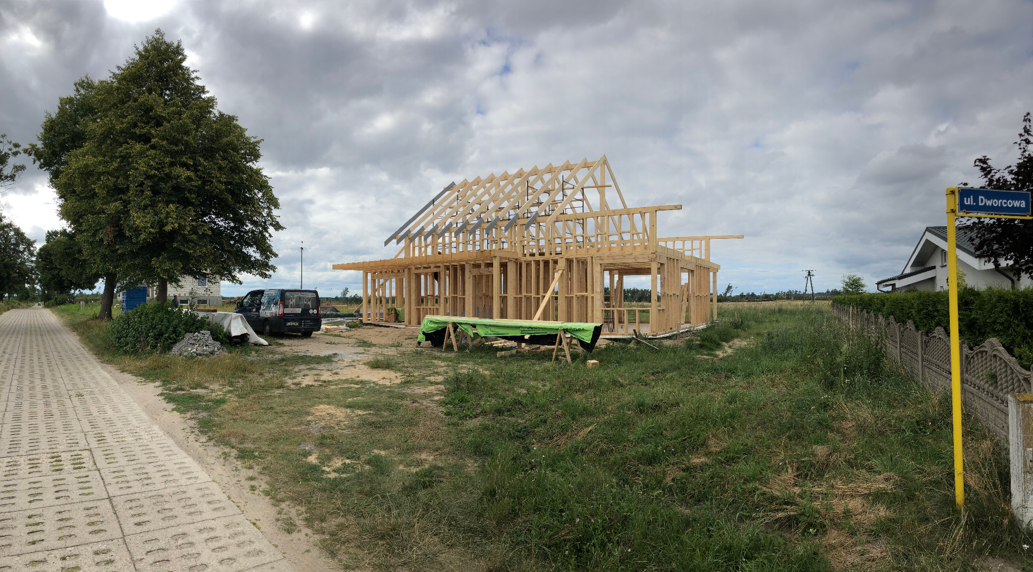 Budowa domu szkieletowego w stanie surowym otwartym, widoczna konstrukcja drewniana, zabezpieczone materiały budowlane, tablica z nazwą ulicy w tle.