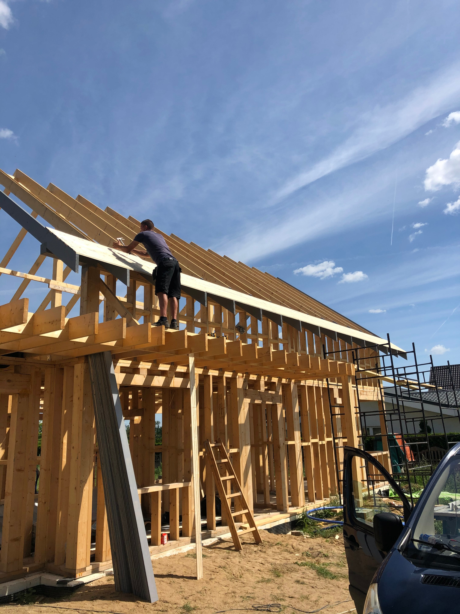 Mężczyzna mocuje płytę do drewnianej konstrukcji dachu domu w trakcie budowy, widok z dołu na tle błękitnego nieba.