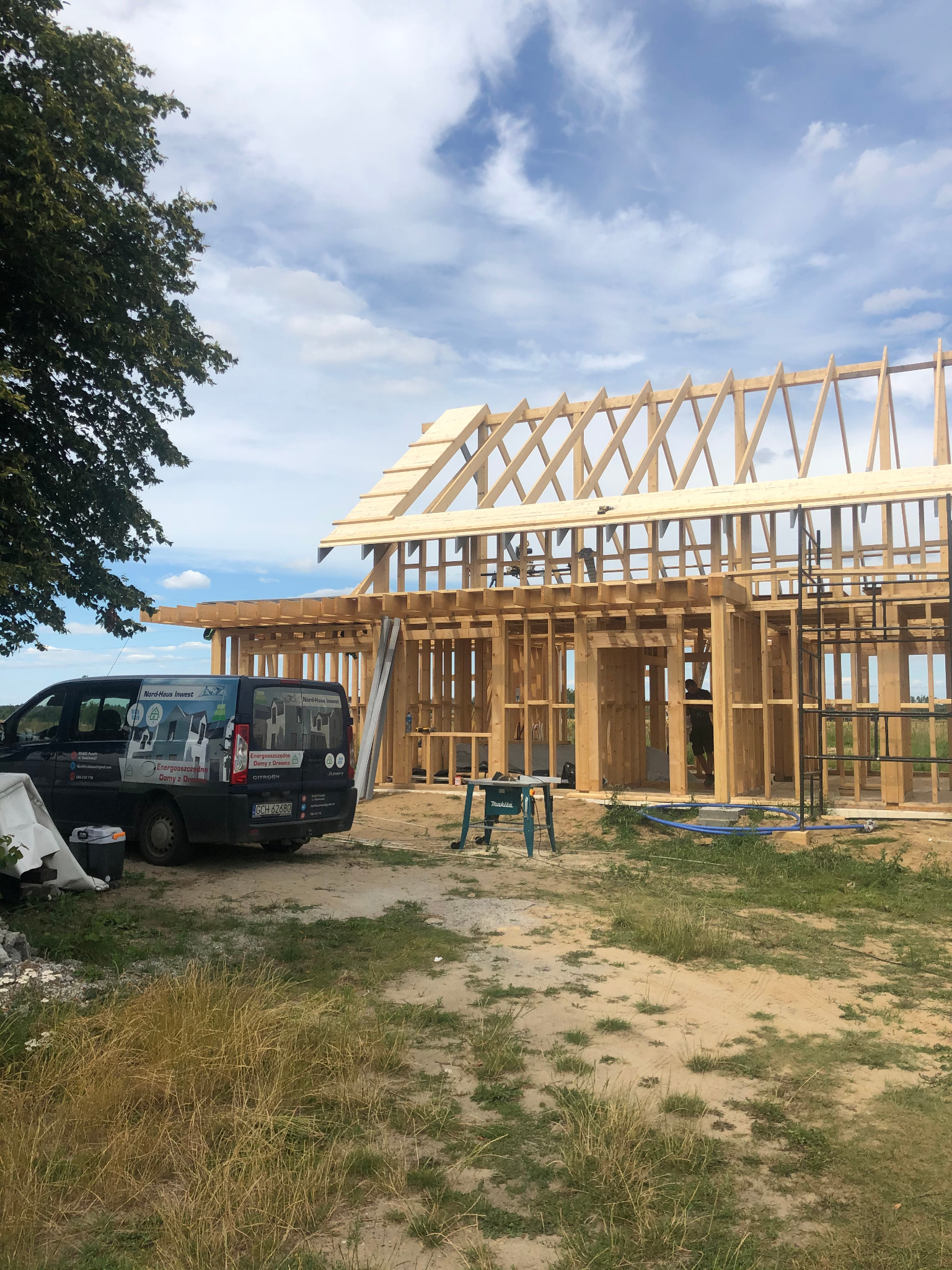 Konstrukcja szkieletowa domu w trakcie budowy, widoczna więźba dachowa, na pierwszym planie samochód dostawczy z reklamą firmy budowlanej.