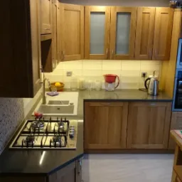 Jasna kuchnia z drewnianymi szafkami, ciemnym blatem i płytą gazową, oświetlona punktowymi lampami sufitowymi i listwą LED pod szafkami wiszącymi.