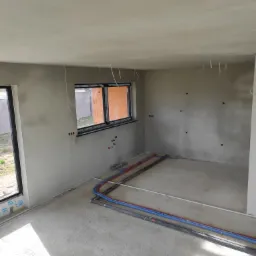 Surowe wnętrze po tynkowaniu maszynowym z widocznymi przewodami instalacji podłogowej; światło słoneczne wpadające przez okno.