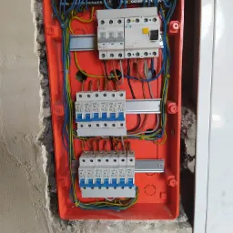 Otwarta, pomarańczowa skrzynka rozdzielcza z zamontowanymi bezpiecznikami i wyłącznikami firmy Eaton i Bemko, widoczne kable w kolorach niebieskim, żółto-zielonym i brązowym, zamocowana na surowej...