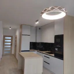 Nowoczesna kuchnia z wyspą, jasne szafki z drewnianymi akcentami, czarny backsplash imitujący marmur, okrągła lampa sufitowa z geometrycznym wzorem, widok na przedpokój z drewnianymi drzwiami.