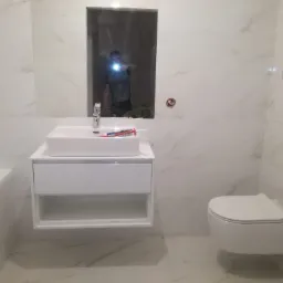 Biała łazienka z marmurowymi płytkami, wiszącą toaletą i umywalką z szafką. Widoczne odbicie osoby w lustrze. Minimalistyczny design, czyste linie i jasne oświetlenie.